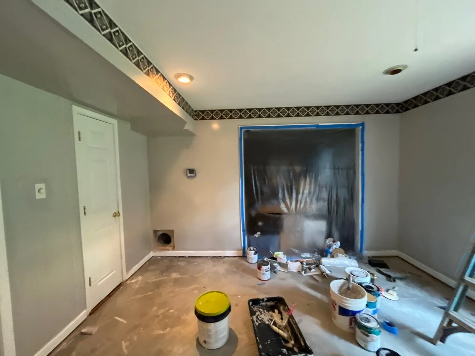 Room under renovation with paint supplies and a window covered with plastic sheeting. Fresh paint and decorative ceiling border visible — serving Dallas, GA.