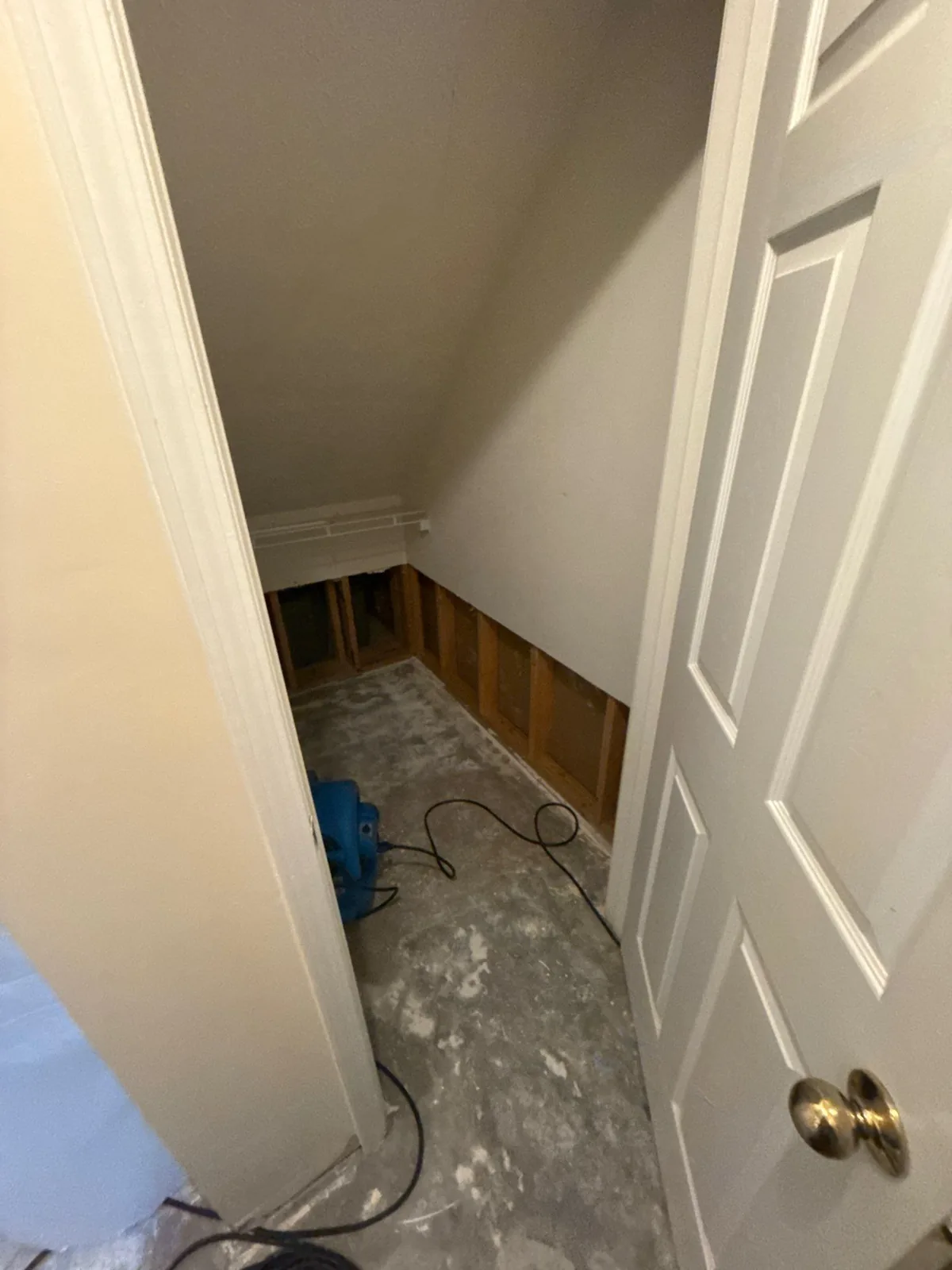Under-stairs area with exposed wall framing and a blue drying fan on the floor, illustrating water mitigation work — serving Dallas, GA.