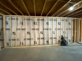 Basement framing phase with Everbilt vapor barrier covering wide stud wall and black trash bag on concrete floor — Metro Atlanta, GA