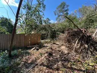 Large uprooted tree with exposed root ball collapsed against new wooden privacy fence in storm-damaged backyard — Acworth, GA