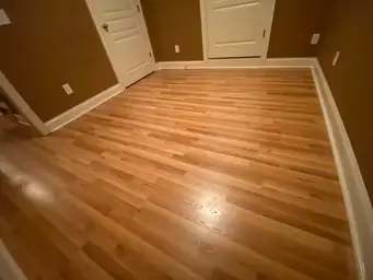 Restored basement room with refinished honey-toned hardwood floors, white baseboard trim, and warm brown walls after water damage cleanup — Marietta, GA